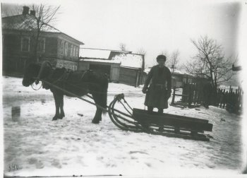 Изображение лошади (слева), запряженной в деревянные сани (справа), за санями мужчина в шапке, теплом шабуре; далее деревья, деревянные дома и постройки, забор; везде снег.