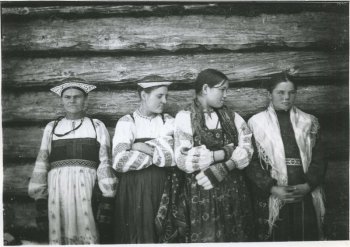 Изображение четырех женщин на фоне бревенчатой стены: на двух женщинах слева кокошники, рубахи с расшитыми на рукавах и у горла узорами, сарафаны, у женщины слева на руках вязаные варежки с узорами; на третьей женщине слева рубаха с расшитыми на рукавах и у горла узорами, сарафан, вязаные перчатки с узорами, на плечах у нее платок; на женщине справа курточка с поясом, юбка, на плечах у нее платок; головы женщин в центре повернуты в бок.