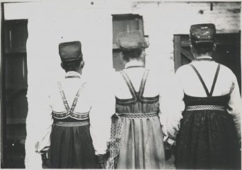 Изображение трех стоящих спиной к объективу женщин в самшурах, белых рубахах, сарафанах с поясами; за женщинами слева видно глухой забор, справа - каменное строение с деревянными воротами. Левее центра полоса стертого изображения (кроме женщины).