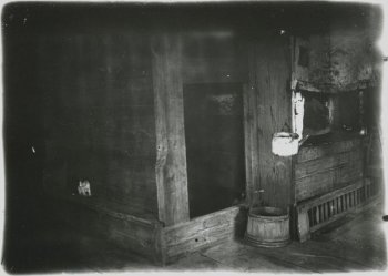Внутренний вид избы: слева в стене деревянная лавка, на ней котенок; в центре полотенце на гвозде, открытая дверь; справа печь, таз для воды, чайник на гвозде, другая хозяйственная утварь.
